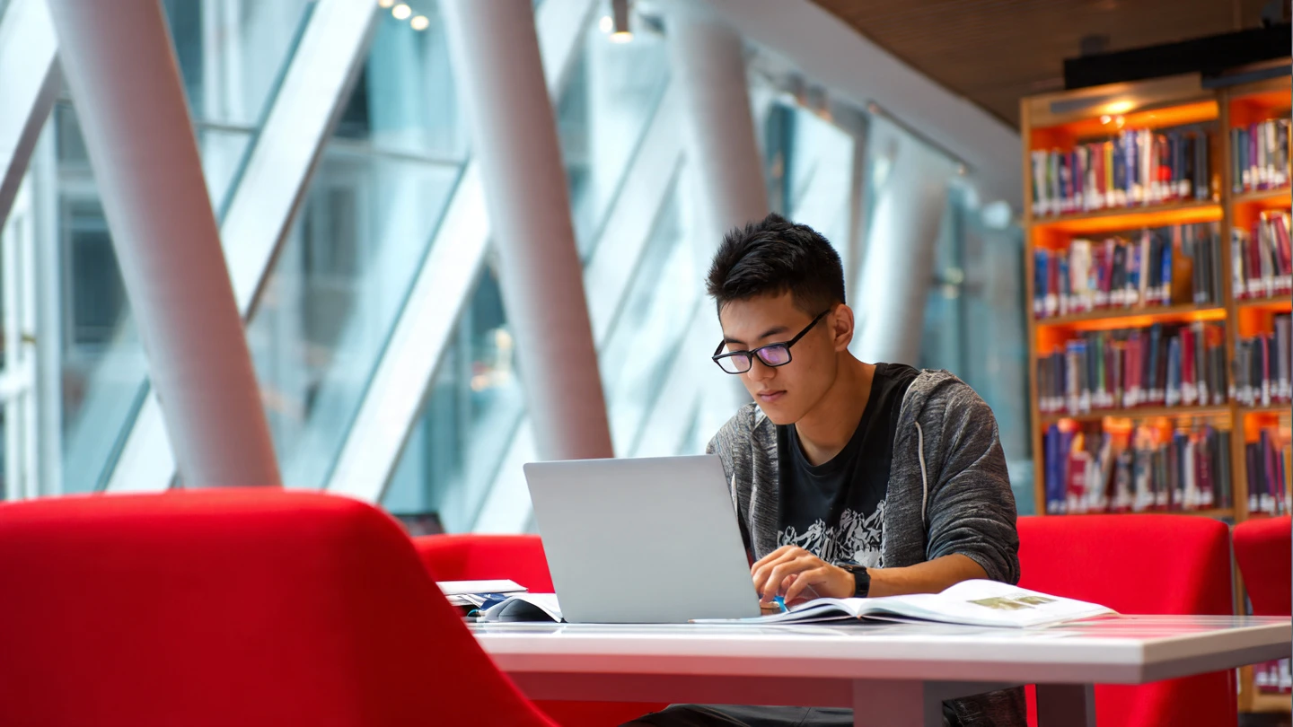 Estudiante joven revisando la beca MEC 2026 requisitos en su portátil en la biblioteca universitaria